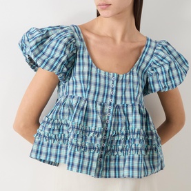 Model wearing Damson Madder blue check cotton blouse with puff sleeves, scoop neckline, button front, and tiered ruched hem detail.