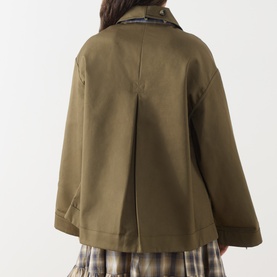 Model wearing Damson Madder olive cotton coat, back view showing pleat detail, styled with a plaid skirt.