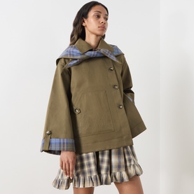 Model wearing Damson Madder olive cotton cape coat with double-breasted buttons and blue check lining, styled with a check mini skirt.