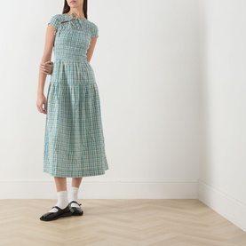 Model wearing Damson Madder multi check cotton midi dress with ruched bodice, bow detail, styled with white socks and black mary jane shoes.