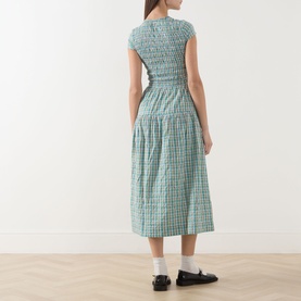 Model wearing Damson Madder dress in multi green gingham check, smocked bodice, midi length, styled with black loafers and socks, rear view.