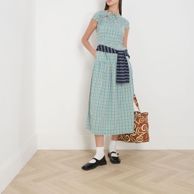 Model wearing Damson Madder multi check cotton midi dress with navy striped sash, styled with white socks, black mary jane shoes and a printed tote