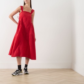 Model wearing Damson Madder red sleeveless midi dress with wide straps and pleated skirt, styled with black socks and chunky trainers.