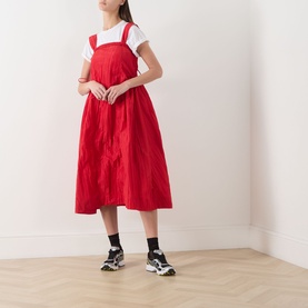 Model wearing Damson Madder red pinafore dress over white T-shirt, paired with black socks and chunky trainers.