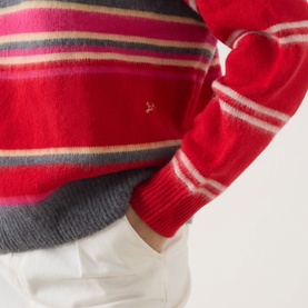 Model wearing Damson Madder pink and red stripe merino wool knit, styled with white trousers. Close-up detail of chest logo and cuffs.