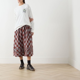 Model wearing Damson Madder Plant Patch Pals T-Shirt in white with long sleeves, styled with red check midi skirt and black trainers.