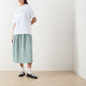 Model wearing Damson Madder white cotton T-shirt, styled with green check midi skirt, white socks and black Mary Jane shoes.