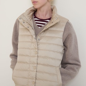 Model wearing Gant Logo Down-Filled Gilet in beige, stand-up collar, snap button placket, styled over a striped top with grey knit sleeves