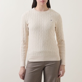 Model wearing Gant Shield Logo Cable Knit Sweater in beige, featuring crew neckline, cable knit texture and long sleeves, styled with taupe trousers.
