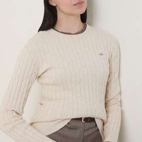 Model wearing Gant Shield Logo Cable Knit Sweater in beige, crew neck, cable knit texture, styled with brown check trousers and belt.