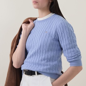 Model wearing Gant Shield Logo Cable Knit Sweater in lavender, crew neck, cable knit detail, styled with white trousers and brown jacket.