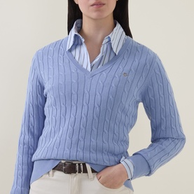 Model wearing Gant Shield Logo Cable Knit Sweater in Misty Lavender, styled over a striped shirt with white jeans.