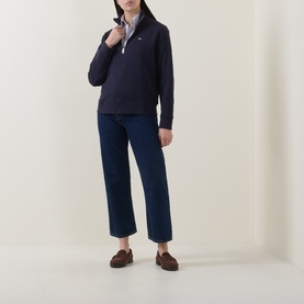 Model wearing Gant Shield Logo Half-Zip Sweatshirt in blue with funnel neck and half-zip. Styled with navy wide-leg jeans and brown loafers.