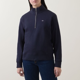Model wearing Gant Shield Logo Half-Zip Sweatshirt in blue, featuring funnel neck, half-zip fastening and small shield logo. Styled with dark jeans.