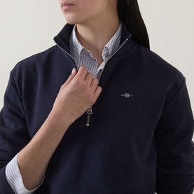 Model wearing Gant Shield Logo Half-Zip Sweatshirt in blue, featuring half-zip funnel neck and embroidered logo, styled over a striped shirt.