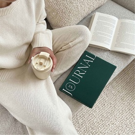 The Head Plan Undated Cloth Covered Journal in Forest Green cloth cover, styled on a sofa beside an open book, with a cream travel cup.