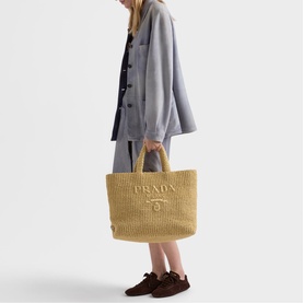 Model carrying Prada Accessories neutral raffia tote bag with embossed logo, styled with grey coat and brown loafers