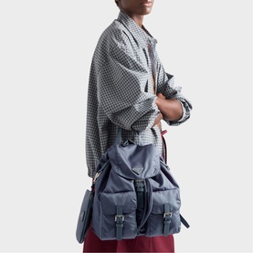 Model carrying Prada Accessories blue fabric backpack with buckle straps, styled with grey checked oversized shirt and burgundy trousers