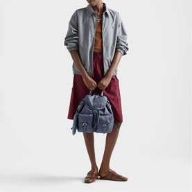 Model carrying Prada Accessories blue fabric backpack with buckle detail, styled with grey shirt, burgundy skirt and tan mules.