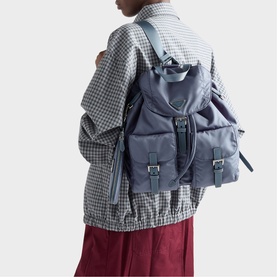 Model carrying Prada Accessories blue nylon backpack with silver buckle straps and front pockets, styled with check jacket and burgundy trousers.
