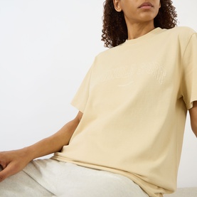 Model wearing Adanola Logo Graphic Oversized T-Shirt in Caramel Beige, round neck, short sleeves, subtle logo detail, styled with light grey trousers.