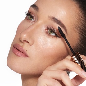 Model with Charlotte Tilbury Eye Shadow in Oyster Twilight, applying mascara wand to lashes, showcasing a soft glam eye look