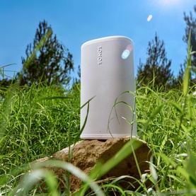 Sonos Play White portable speaker in white, placed on a rock outdoors surrounded by green grass under a blue sky.