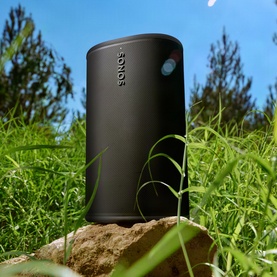 Sonos Play Black portable speaker in black, positioned outdoors on a rock surrounded by green grass under a blue sky.