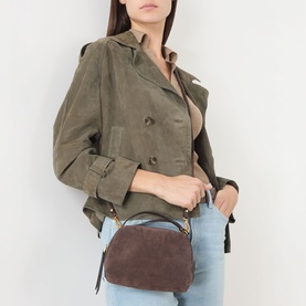 Model carrying Gianni Chiarini Alifa Crossbody Bag in brown suede with gold-tone hardware, worn crossbody, styled with olive jacket and jeans.