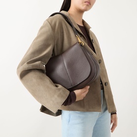 Model carrying Gianni Chiarini Helena Crossbody Bag in brown grained leather with gold-tone hardware, styled with a beige suede jacket and light blue