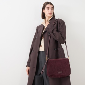 Model carrying Gianni Chiarini Nora Crossbody Bag in burgundy suede with gold-tone hardware, worn crossbody, styled with dark trench coat.