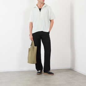 Model wearing Issey Miyake Duomo Green short-sleeve casual shirt with black trousers, carrying an olive tote bag, wearing black loafers.