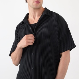 Model wearing Issey Miyake black pleated short-sleeve casual shirt with textured ribbed finish, styled over a black crew-neck top.