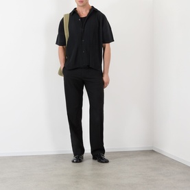 Model wearing Issey Miyake black short-sleeve casual shirt with black trousers and khaki cross-body bag, black loafers.