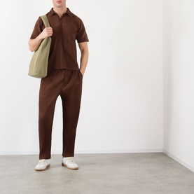 Model wearing Issey Miyake Anchor Brown casual trousers with matching brown polo shirt, white trainers, and carrying a sage green tote bag.