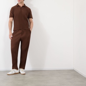 Model wearing Issey Miyake anchor brown short-sleeve polo shirt with open collar, styled with matching brown trousers and white trainers.