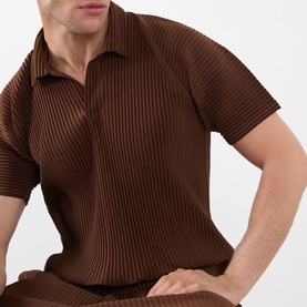 Model wearing Issey Miyake anchor brown pleated polo shirt with open collar, styled with matching pleated trousers.
