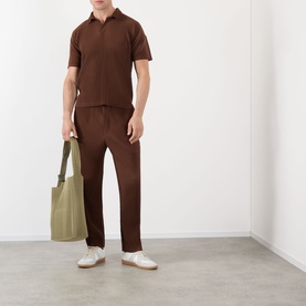 Model wearing Issey Miyake Anchor Brown polo shirt and matching trousers, styled with white trainers and carrying an olive tote bag.