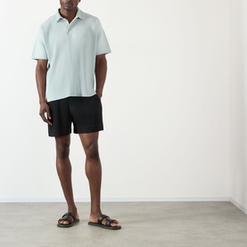 Model wearing Issey Miyake polo in Boboli Moss Ash, styled with black shorts and brown leather sandals.
