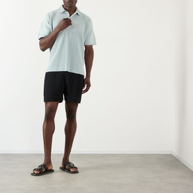 Model wearing Issey Miyake polo in Boboli Moss Ash, styled with black shorts and brown leather slide sandals