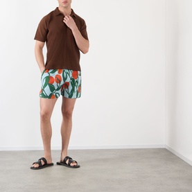 Model wearing Issey Miyake blue fruit-print swim shorts with a brown polo shirt and black sandals.