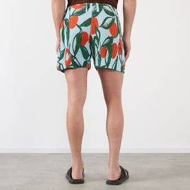 Model wearing Issey Miyake blue polyester shorts with orange and green tropical print, paired with black sandals, rear view.