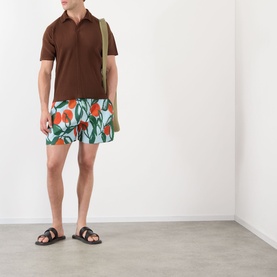 Model wearing Issey Miyake blue floral print swim shorts, styled with a brown polo shirt and dark slide sandals, carrying an olive tote bag.