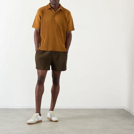 Model wearing Issey Miyake Genova Metal Ocher shorts in olive-brown, paired with a tan polo shirt and white trainers.