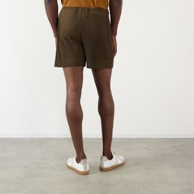 Model wearing Issey Miyake shorts in Genova Metal Ocher, pleated texture, styled with white low-top trainers, rear view.