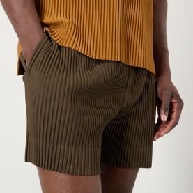 Model wearing Issey Miyake pleated shorts in Genova Metal Ocher, styled with a mustard pleated top. Side pocket detail visible.