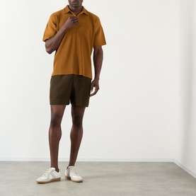 Model wearing Issey Miyake Shorts in Genova Metal Ocher, styled with a mustard polo shirt and white trainers.