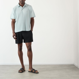 Model wearing Issey Miyake black polyester shorts with a light blue polo shirt and brown double-strap sandals.