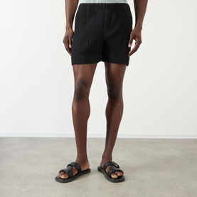 Model wearing Issey Miyake black pleated polyester shorts, styled with black leather slide sandals.