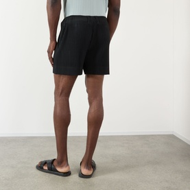 Model wearing Issey Miyake black pleated polyester shorts, styled with black leather sandals, seen from behind.